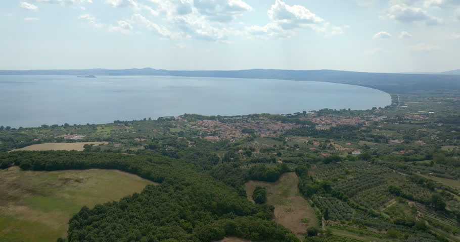 Panoramic aerial view of a Bolsena Lake, in Lazio, Italy, surrounded by hills, with a small town on its shore and countryside with olive groves, woods and scattered farmhouses under a cloudy sky.