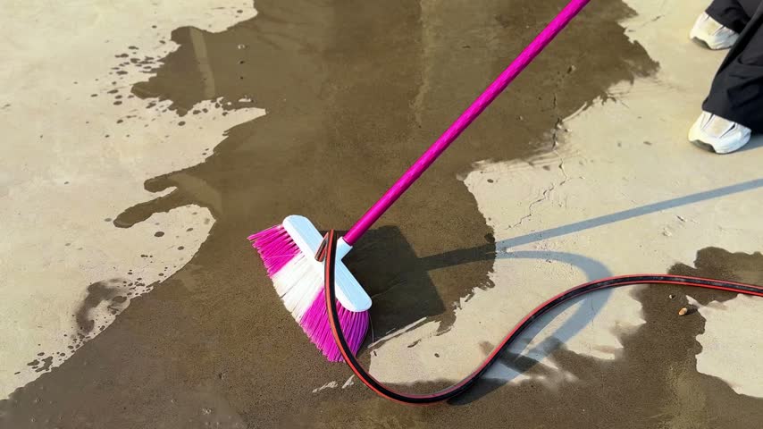 A person wearing black pants sweeps a wet outdoor concrete floor using a pink push broom and a running water hose during bright daylight. Strong shadows reflect across the surface.