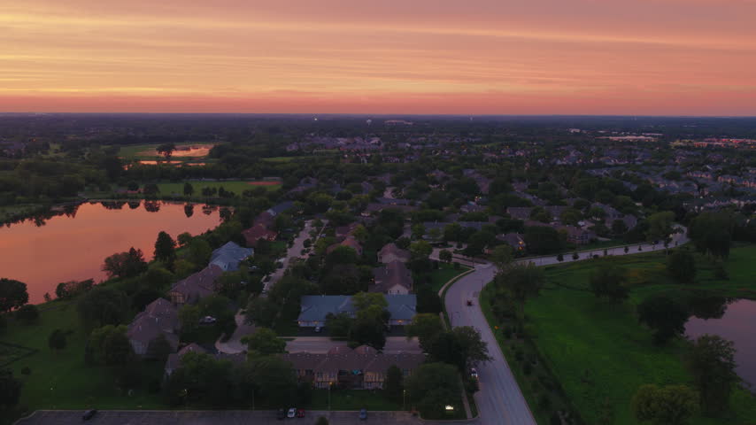 Century Park Arboretum in Vernon Hills, Illinois, homes and tree lined streets beside a still pond under a warm orange evening sky.