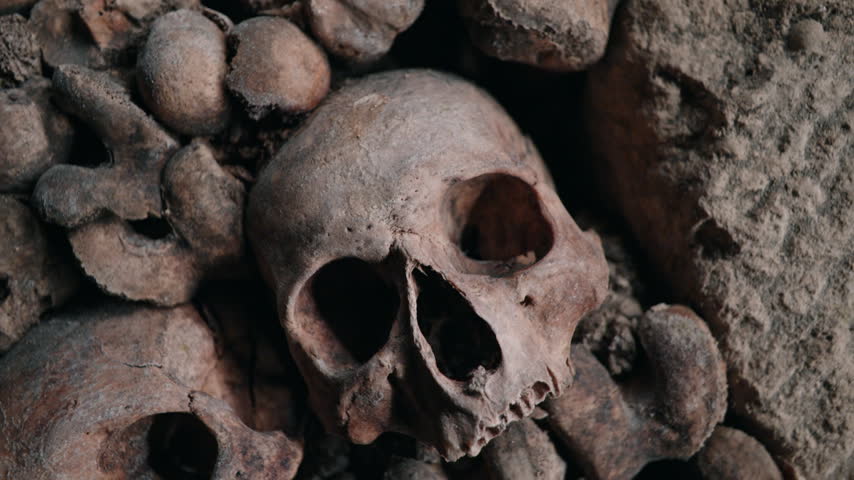 Tight shot lingers on several worn craniums packed among rough bones, creating a dark, somber burial backdrop.