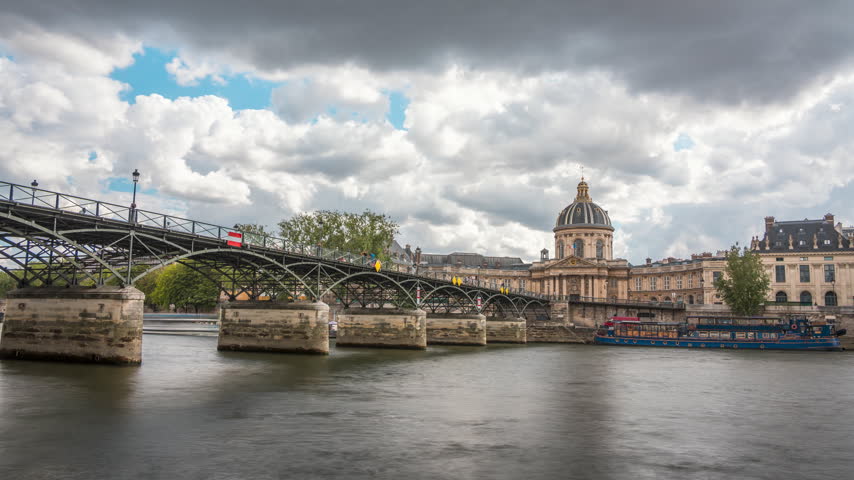 Boats move along the Seine beneath Pont des Arts as the Institut de France rises under fast-moving clouds.