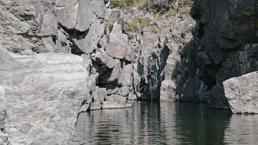 Steep grey rock walls rise above a calm water channel in a narrow gorge under clear daylight.