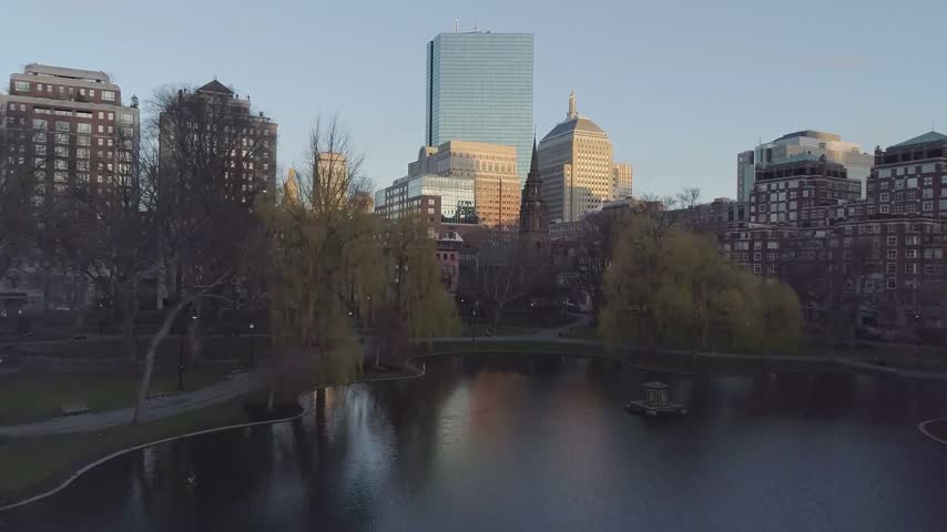 A beautiful high-angle drone perspective looking from the edge of the Boston Public Garden toward the city center skyline of Boston, Massachusetts. The footage highlights the towering blue glass facade of the John Hancock Tower rising above a dense mix of historic brick residential blocks, hotel buildings, and urban park trees under the soft light of a clear morning sky.