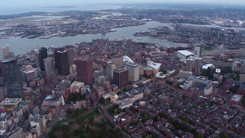 A beautiful wide, high-angle drone panorama looking over the tightly packed brick townhouses of the Beacon Hill neighborhood and the Massachusetts State House in Boston, Massachusetts. The footage captures the historic city streets transitioning into the downtown financial district skyscrapers, with the Boston Inner Harbor, East Boston, and Logan International Airport visible in the background during the evening twilight.