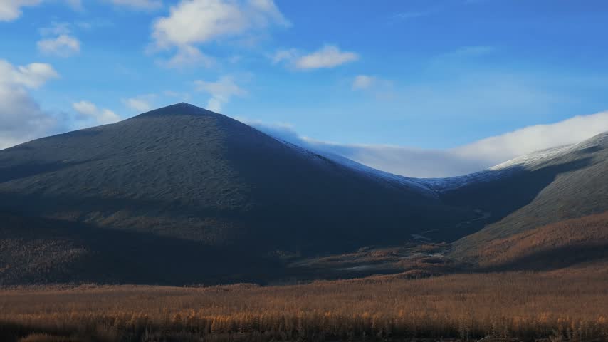 Snow-capped mountain peaks rise above a dense golden-brown forest and distant winding river, with drifting clouds casting shifting shadows across the vast landscape under a bright blue sky.