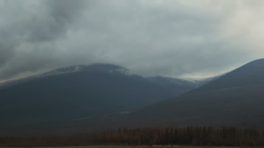 Time-lapse of clouds drifting left to right across snow-capped mountain peaks, partially obscuring the summits while brownish trees remain static in the foreground under a dim, overcast sky.