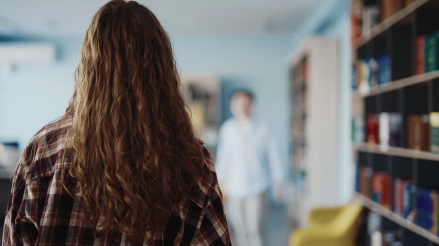 A woman with long curly hair in a plaid shirt walks toward a man in a library, raising her hand to give him a high five. They connect hands with a smile, sharing a moment of joy and camaraderie in a