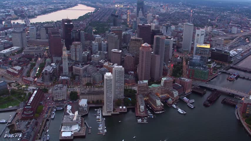 A beautiful high-angle panoramic drone shot looking over the dense urban architecture of downtown Boston, Massachusetts, during the evening twilight. The footage captures the illuminated high-rises and corporate skyscrapers of the financial district, with the historic Custom House Tower visible on the left, leading toward the calm waters of the Charles River winding through the background under a moody evening sky.
