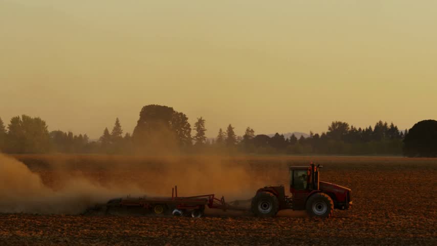 Tractor plowing agricultural farm field at sunset with dust rising