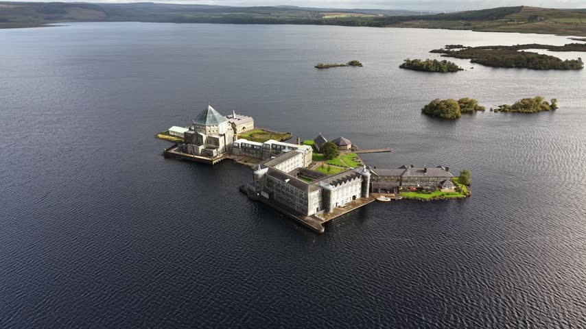 Lough Derg, County Donegal, Ireland — 5 October 2026. Close aerial drone footage approaching Station Island, home to St Patrick’s Purgatory, one of Ireland’s historic Christian pilgrimage sites. The view highlights St Patrick’s Basilica and the adjoining sanctuary buildings surrounded by the dark waters of Lough Derg, with small wooded islands, distant wind turbines and dramatic cloud formations across the surrounding countryside.