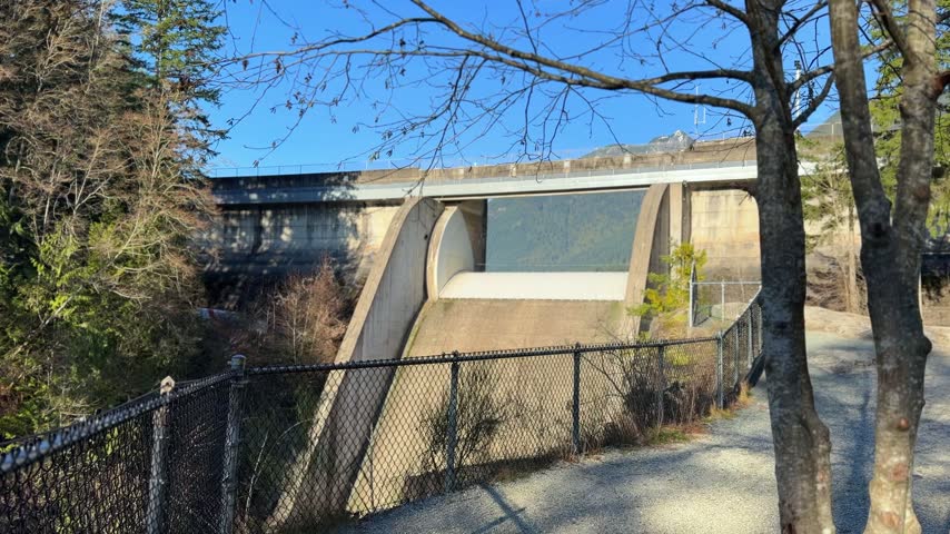 Concrete arch and spillway of Cleveland Dam at Capilano Lake in Vancouver on a sunny winter day, with the dam structure, mountains and blue sky behind the trees.