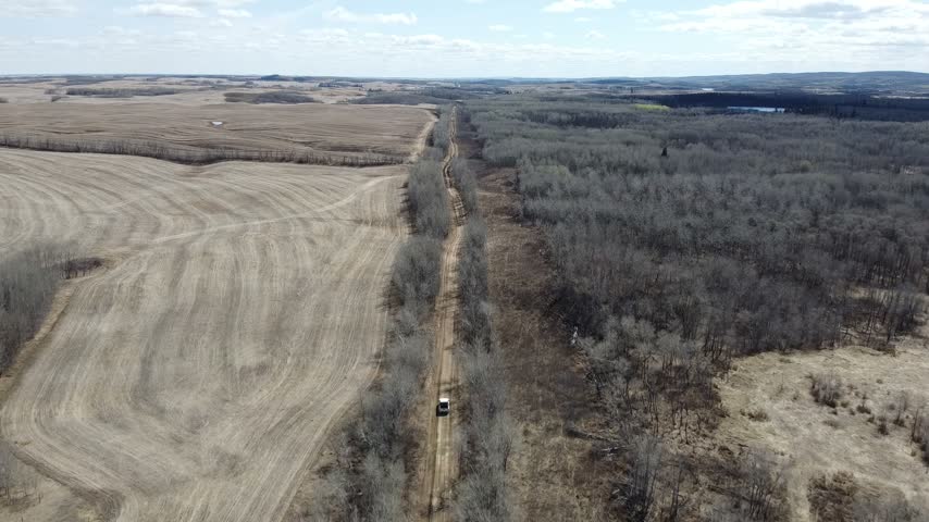 drone shot following a pickup truck driving along a long, straight dirt road dividing harvested agricultural fields and a dense forest