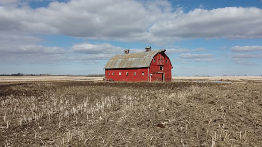 An aerial drone shot captures a lonely red barn standing in the middle of a vast, flat agricultural field with standing water puddles