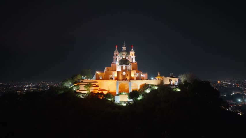 Aerial view of the illuminated Santuario de la Virgen de los Remedios perched on a hill at night, overlooking the city lights of Cholula, Puebla, Mexico.