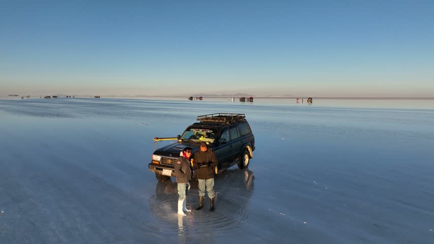 Aerial view of Uyuni Salt Flat with a parked off-road vehicle and two people standing on the reflective surface under a clear sky in Potosi Department, Bolivia.
