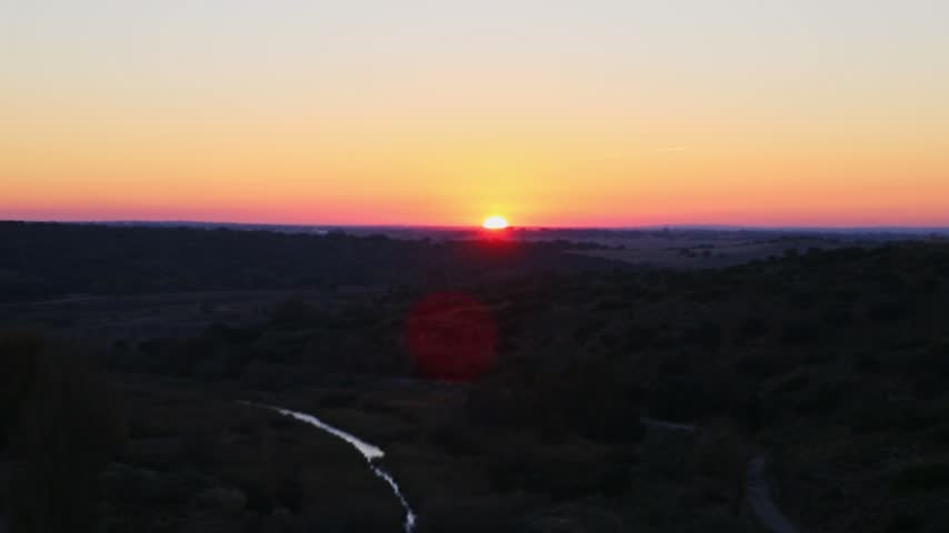An aerial pan shot starting with a bright sunset over the surrounding landscape and valley, panning left to reveal the overflowing Peñarroya dam face and spillway in Ruidera.