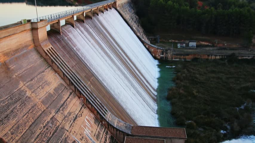 An aerial pan shot starting with a bright sunset over the surrounding landscape and valley, panning left to reveal the overflowing Peñarroya dam face and spillway in Ruidera.
