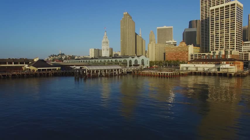 A beautiful high-angle panoramic drone shot capturing the historic San Francisco Ferry Building and its clock tower along the Embarcadero waterfront in San Francisco, California. The footage highlights the calm waters of the bay and historic piers in the foreground, leading toward a dense cluster of financial district skyscrapers, including the Transamerica Pyramid and modern corporate high-rises, illuminated by the warm light of a clear early morning sky.
