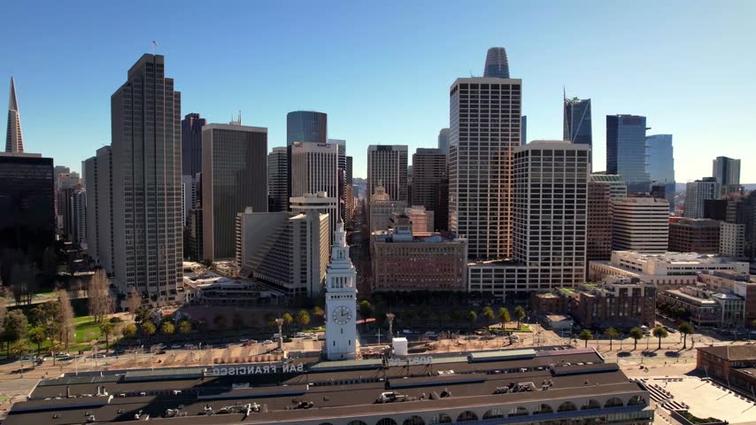 A beautiful high-angle panoramic drone shot looking over the urban plaza surrounding the historic San Francisco Ferry Building in San Francisco, California. The footage highlights the architectural details of the historic clock tower in the right foreground, leading the eye across Sue Bierman Park and the Embarcadero toward the massive corporate skyscrapers of the financial district—including the iconic Transamerica Pyramid and the towering Salesforce Tower—standing under a bright, clear blue morning sky.
