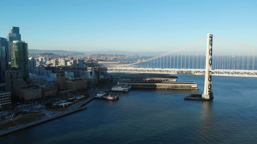 A beautiful high-angle panoramic drone shot looking down at the massive white suspension tower and steel truss architecture of the San Francisco–Oakland Bay Bridge in San Francisco, California. The footage captures vehicle traffic crossing the upper deck, shaded waterfront piers along the Embarcadero, and the sweeping urban cityscape stretching toward distant coastal hills under a crisp, clear blue morning sky.
