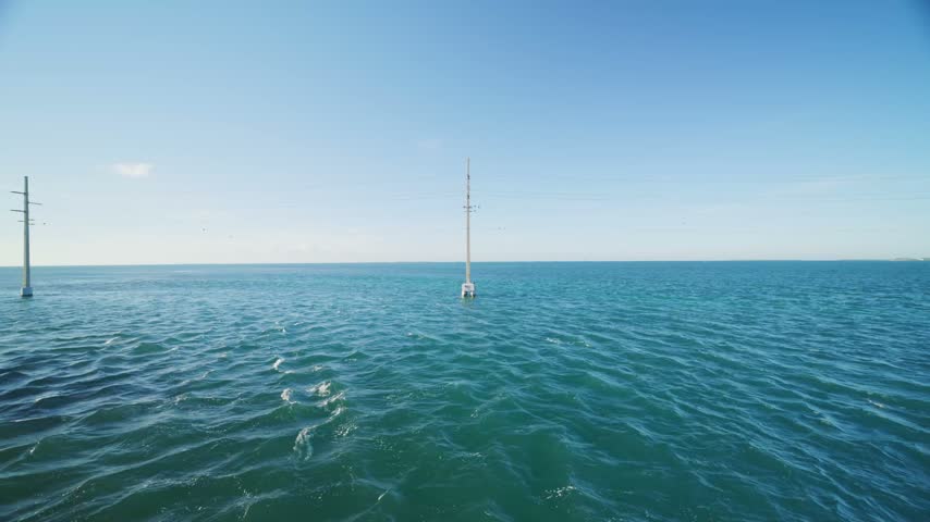 Utility poles stand in the blue seawater. Power cables cross the clear sky. Florida Keys coastal landscape. Key West travel destination.