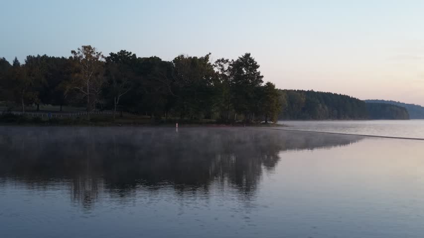Early morning fog drifts over a calm lake in the foreground, with a tree-lined shoreline fading into soft mist in the background at dawn.