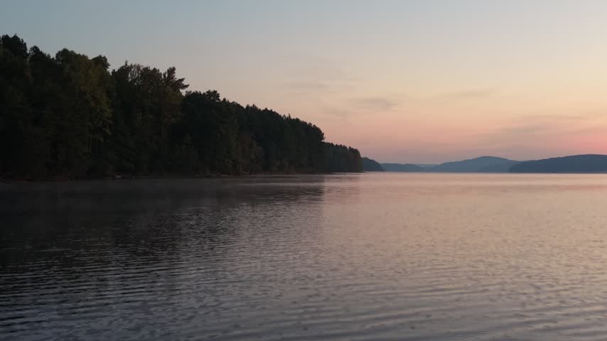 A dense forested shoreline with touches of early autumn color stands over dark calm water, soft reflections under a pale overcast sky.