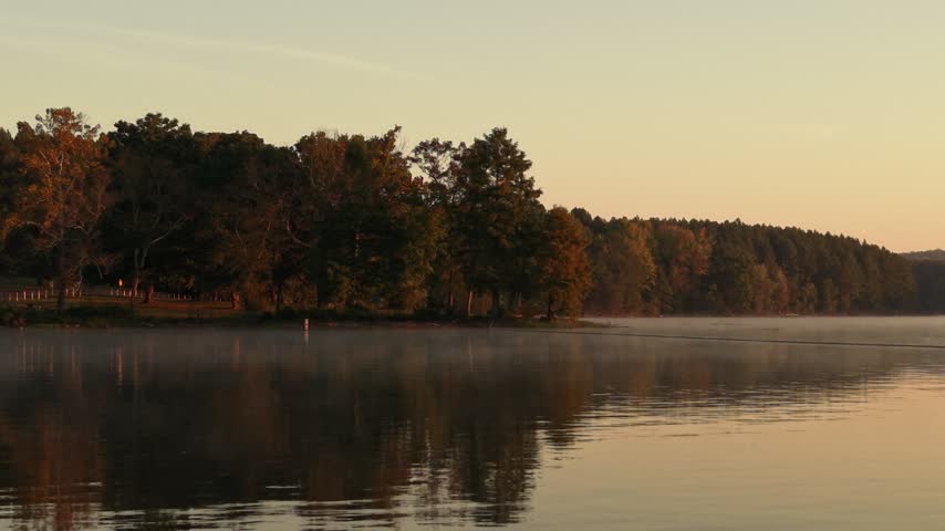 Early morning fog drifts over a calm lake in the foreground, with a tree-lined shoreline fading into soft mist in the background at dawn.