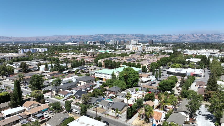 San Jose, California, USA - Drone footage overlooking the downtown district with residential areas and technology office buildings extending across the city. The view highlights the urban skyline, Silicon Valley development, surrounding neighborhoods, and distant East Bay mountains under clear sunny conditions.