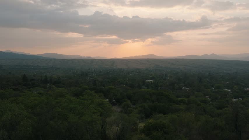 Soft amber light breaks through clouds above a distant mountain range as dusk settles over a vast green forest canopy.