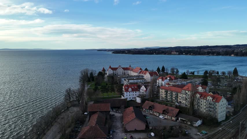 Aerial view past white waterfront buildings with red roofs toward a broad lake under a cloudy sky.