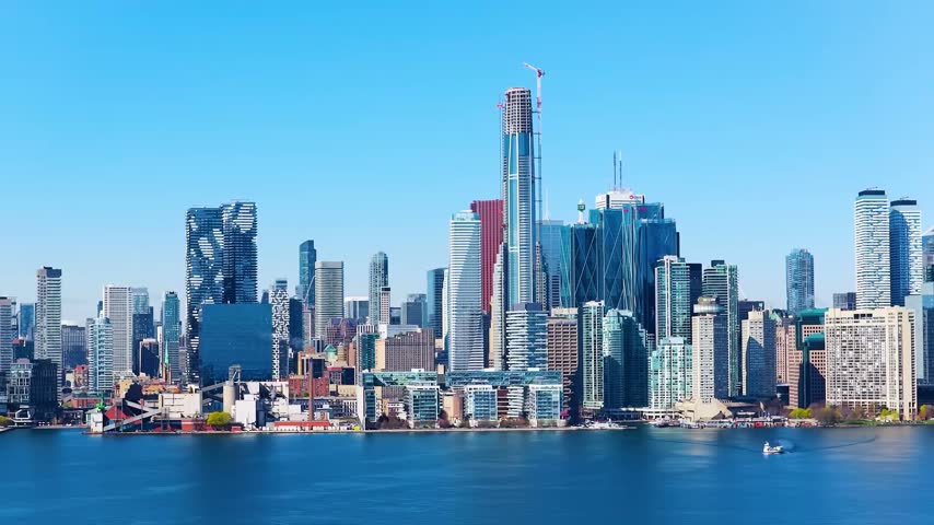 Wide panoramic look across Lake Ontario at the downtown Toronto, Ontario, Canada city skyline on a clear sunny day, showcasing the distinct curved facade of Aura and Pinnacle One Yonge (SkyTower) under construction with massive tower cranes.