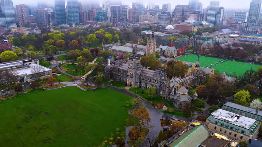 Wide aerial drone shot of the historic University College Romanesque Revival building on the University of Toronto St. George Campus in Toronto, Ontario, Canada, showcasing the grand stone facade, wet paths, Front Campus green lawn, and a misty downtown skyline.