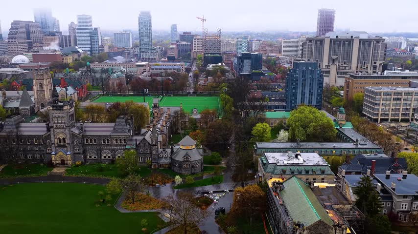 Aerial drone shot of the historic University College building on the University of Toronto St. George Campus in Toronto, Ontario, Canada, featuring its Romanesque Revival architecture, the vast Front Campus green lawn, and the downtown city skyline shrouded in fog.