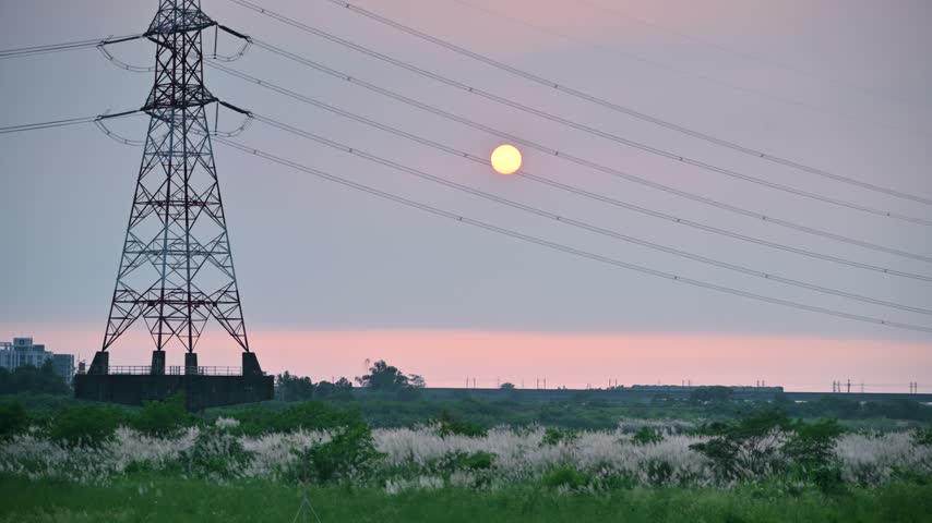 High voltage power tower standing near grassy fields during a peaceful autumn sunset in Tainan, Taiwan.