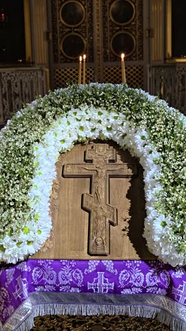 A wooden cross adorned with white flowers and wrapped in a purple cloth.
