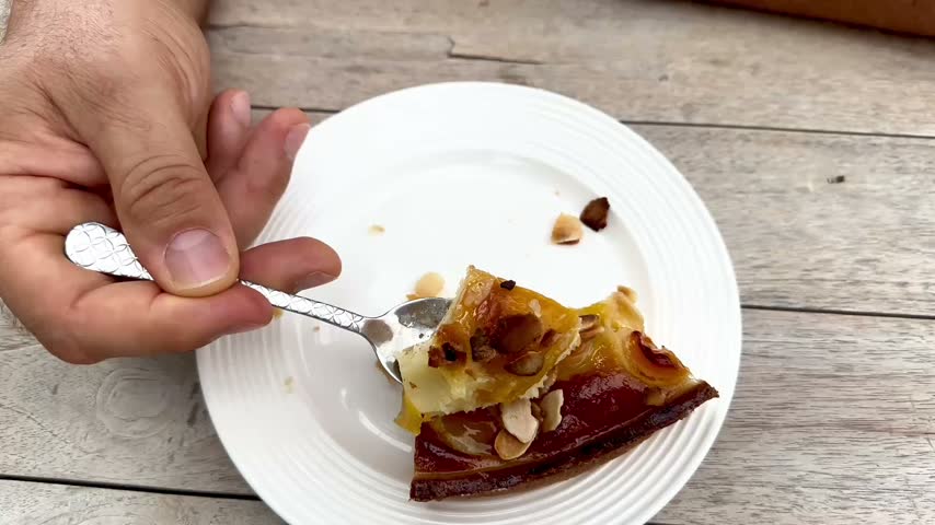 Close-up shot of a person using a spoon to eat a delicious bite of homemade apricot and almond tart from a white plate on a wooden table outdoors.