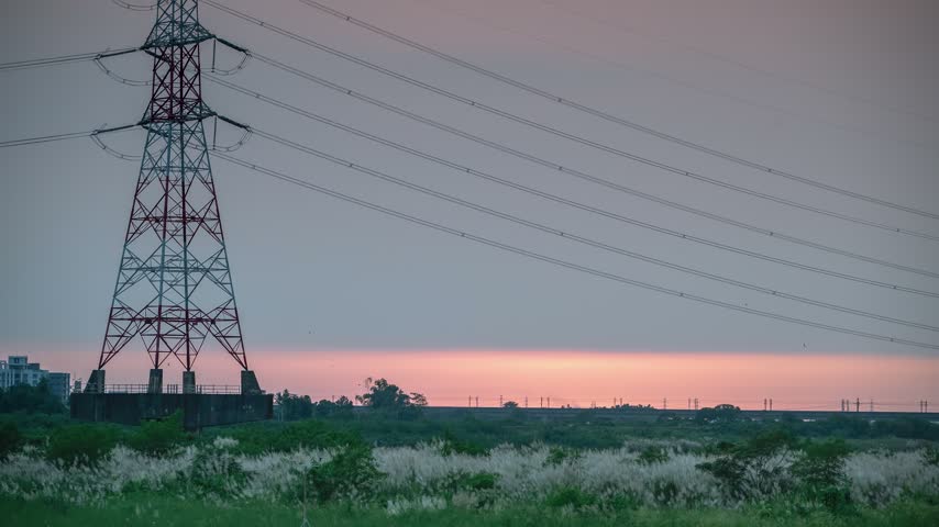 High voltage electricity tower standing near a riverbank covered with white grass during a vibrant sunset in Tainan, Taiwan.