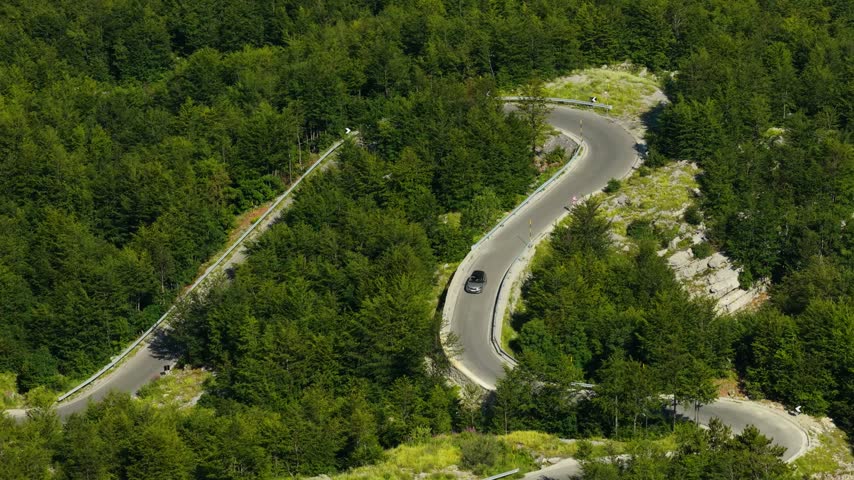 Aerial view of a dark sedan driving around a series of hairpin bends on a steep rocky road through dense forest in Albanian Alps National Park near Shale Albania. The location is in the Prokletije, also known as the Accursed Mountains or Albanian Alps.
