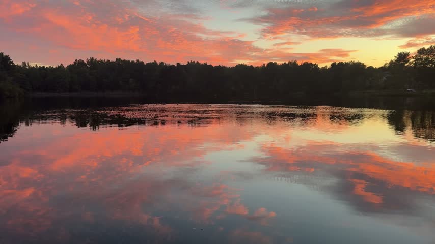Colorful sunset clouds reflected across a calm lake surrounded by a dark tree-lined shoreline