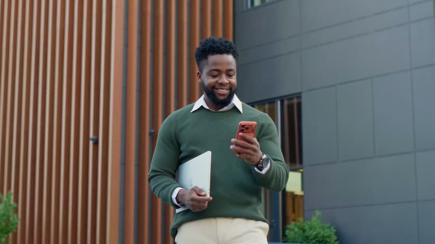 Confident young businessman walking outdoors carrying a laptop and using a mobile phone