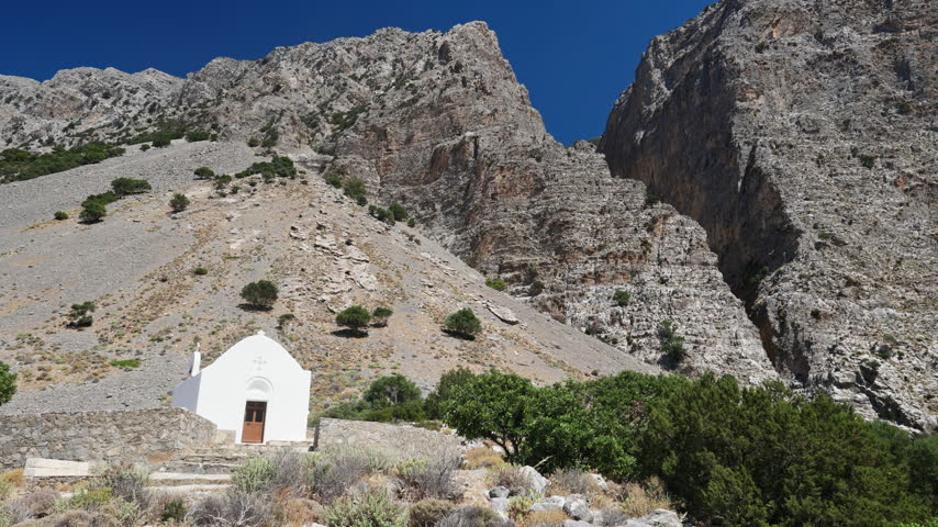 Traditional white Greek Orthodox chapel situated at the entrance of the towering Ha Gorge in Crete, Greece, rocky cliffs rising into a blue sky