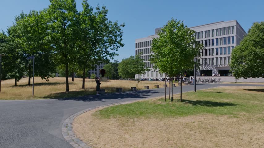 Wide goethe university campus with modern building, curved walkway, trees, dry grass, bicycle racks, and one distant person under clear blue sky showing calm academic outdoor setting