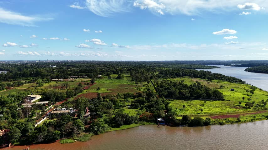 Aerial drone view over a wide lake surrounded by lush green countryside, farmland, rural properties and dense vegetation, with small boats navigating near the shoreline under a blue sky.