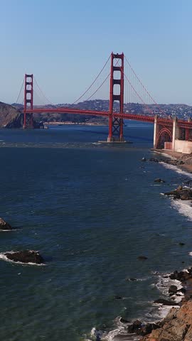 Aerial perspective reveals the Golden Gate Bridge connecting the headlands across the bay, while ocean waves wash onto Baker Beach along the rugged San Francisco coastline on a clear sunny day.