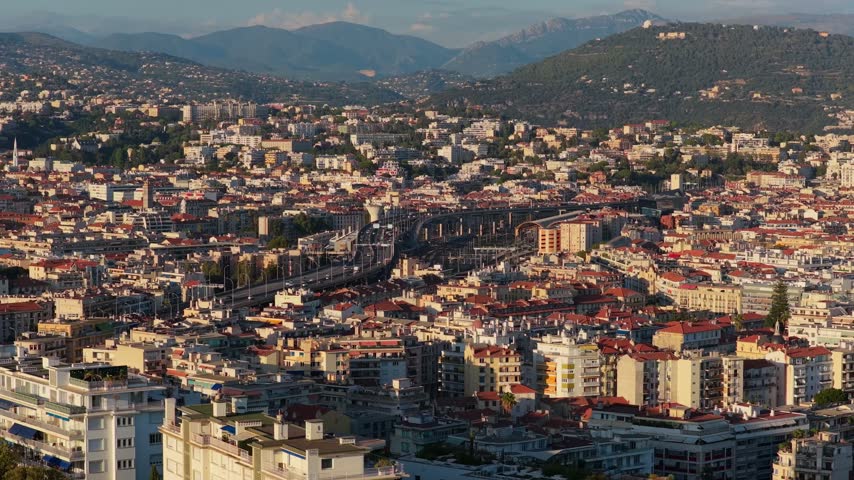 Static telephoto of dense rooftops and an elevated expressway curving past the rail station to green hills, evening light, Nice, French Riviera, Cote d Azur, France