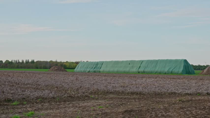 Wide panoramic view of an agricultural field with a large silage storage covered with a green plastic tarp in a rural landscape under a cloudy sky