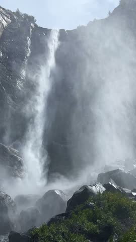 Powerful Bridalveil Fall cascades down a steep granite cliff, creating swirling mist and spray above large boulders in Yosemite National Park, California, USA.