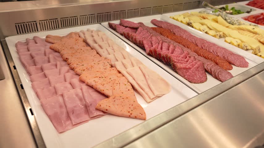 Sliced cold cuts and sausages are arranged in a chilled buffet counter. Cheese and vegetables are displayed beside meats.