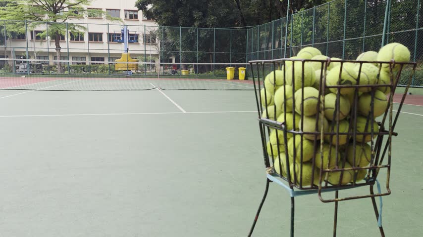 a green tennis court, featuring a wire metal basket filled with piles of yellow tennis balls in the lower right foreground.